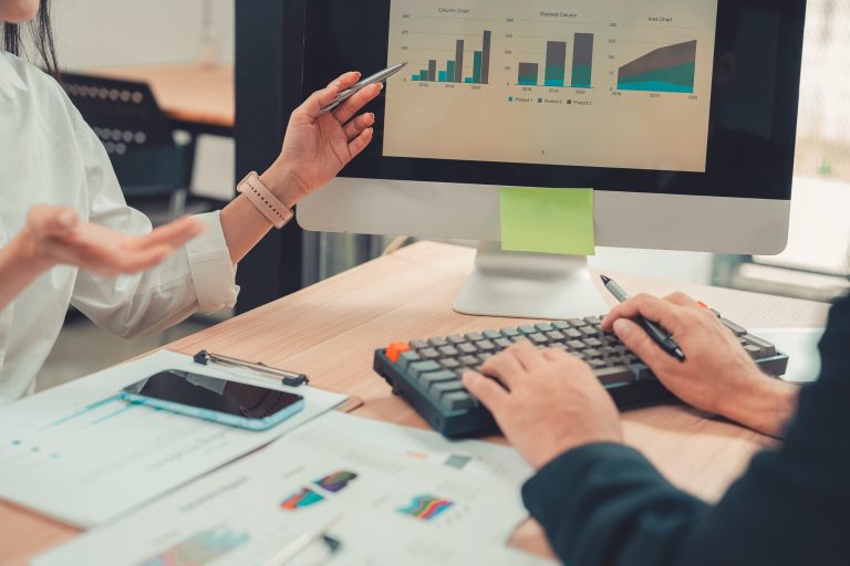 Two accountants use calculators to perform company financial calculations, handle accounting on laptop computers at their desks, manage budgets, and handle financial tasks.